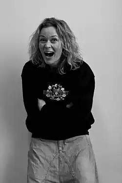 black-and-white image of Line Verndal wearing a dark patterned sweater and denim pants, slightly hunched forward with crossed arms, laughing broadly at camera