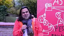 this person wearing a pink coat and a scarf they have long dark hair and a thin white beard