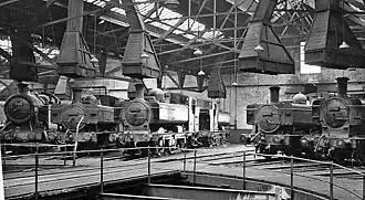 The inside of an engine shed with part of a turntable in the foreground. A number of engines are arranged round the turntable, each with their chimneys under large ventilation cowls.