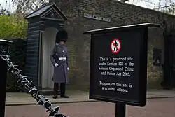 A wall with the street sign "Stable Yard Road", in front of which stands an armed guard wearing a bearskin cap. A free-standing sign in the foreground reads: This is a protected site under Section 128 of the Serious Organised Crime and Police Act 2005. Trespass on this site is a criminal offence.