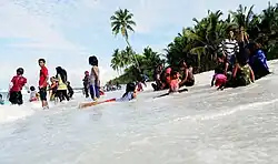 A photo of dark-skinned boys and girls standing, sitting and lying in wet clothes on the surf of a beach. Some palm trees stand in the background. Some women in veils are among the kids.