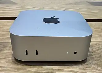 A Mac mini computer (shaped like a small aluminum box) is pictured on top of a wooden table.