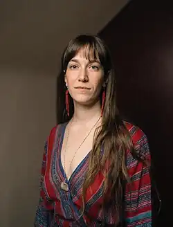 Madeleine has long brown hair with bangs. She is wearing a red and blue striped shirt.