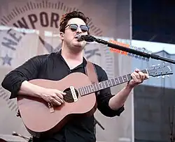 Marcus Mumford, in sunglasses and a black shirt, playing a guitar and singing onto a mic