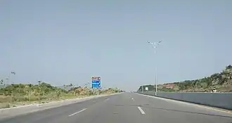 This is a picture of Margalla Avenue near Shah Allah Ditta. It shows a three east-bound lanes of a road in the foreground. It is lined with plants and bushes on either side. There is a blueish sky in the background. Other features present are a signboard on the centre-left, and a street light on the centre-right, either side of the road.