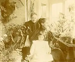 Two women in formal dress (one seated, the other standing behind the first) in a room filled with plants. One wall is all windows with bright sunlight shining in. In front of the two women is a small child, who appears to be about 6 years old, in a white dress.