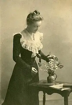 Woman in formal black dress with a white ruffle standing by a small table on which rests a vase of flowers and a few books. The woman is looking intently at the flowers and touching them with one hand, as if arranging them