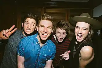 Four men are shown in an upper body shot. They are all slightly crouched and leaning towards the viewer. The man at right has dark hair with his right eye almost closed and holds his right hand up with a V-for-victory gesture. The second man has light brown hair and a wide smile. The third man has brown hair and holds his left hand with a thumb up. The last man wears a hat over dark longer hair and winks with his left eye.