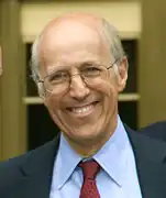 Caucasian exhibiting male-pattern baldness smiling; wearing spectacles, a blue suit and red tie