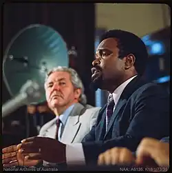 Michael Somare sitting at a desk speaking with an out of focus microphone in front of the shot