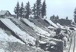 Men working next to chords of wood at the bottom of wooden ramps on the side of a hill. A building and set of trees is on top of the hill. A rail line parallels the chords of wood.
