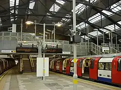A train, with its red sliding doors open, waits at the right side of a platform. Steps lead up from the platform to a metal bridge crossing the tracks and a partly glazed roof arches on steel trusses high above.