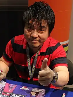 Producer Motohide Eshiro looks at the camera and gives a thumbs up at the 2016 Anime Expo, where he promoted the sixth mainline Ace Attorney installment, Spirit of Justice