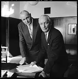 Two men in suits and ties standing by a desk looking at paper work. Mr E C Fussellon the right as retiring Governor of Reserve Bank, with his successor, Mr G Wilson on the left.