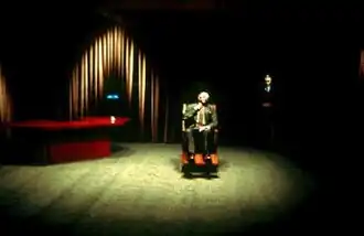 A short, strange-looking man seated in a large wooden wheelchair under an intense beam of light in a large and sparsely furnished room; a desk is in a far corner and the walls are covered in curtains.