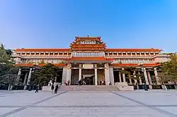 An ornate building front stretching the width of the image, slightly tilted, against a uniformly blue sky. Its front has a projecting pavilion in the Chinese style, echoed by a similar pagoda-style top on the roof above it.