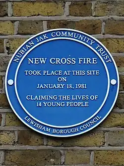 A blue circular plaque sits on the exterior of a brick building’s wall. The plaque reads “New Cross Fire took place on this site on January 18, 1981 claiming the lives of 14 young people.” The outer ring of the plaque reads “Nubian Jak Community Trust; Lewisham Borough Council”