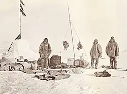 On the left is a snow cairn with flags. Three men are nearby, and assorted equipment is strewn on the snow.