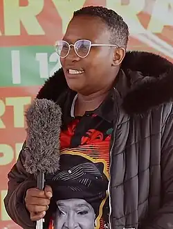 A woman standing and holding a microphone. She is wearing glasses, a large black puffer jacket, and a black polo shirt