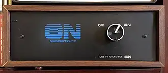 A front view of a wooden box with wood-veneer exterior and a black front. A blue "ON Subscription TV" logo, with the ON letters in a linear gradient, graces the front. A silver knob selects between two options: OFF and ON (stylized like the logo). Beneath the knob is the instruction "TUNE TV TO CH 3 FOR ON".