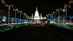 The temple grounds and Oakland California Temple at night during the Christmas season, with several plants and trees covered in Christmas lights.