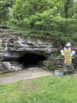 Paved entrance to cave, a opening that is wider than it is tall, in stone that is covered with vines and other plants. The current welcome sign and a container of flowers sits to the right of the entrance.