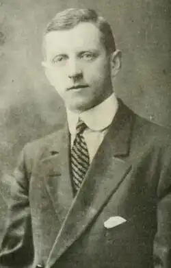 Black and white photo of a man wearing a dark suit jacket, a high-collared white dress shirt, and a striped necktie