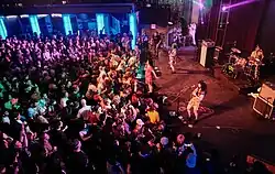 The band Otoboke Beaver performs on a stage illuminated by vibrant multicolored lights at The Palace Theatre during Sled Island 2025. The group includes a vocalist, drummer, guitarist, and bassist, all actively engaged in the music. A large crowd fills the foreground, many with hands raised, enjoying the lively concert. Vertical blue lights frame the stage, and a balcony area with additional spectators adds depth to the venue’s energetic ambiance.