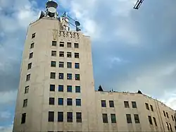 Ground-level view of a 10-story building; the exterior has a tan hue with dark windows and a higher front side