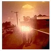 A sepia-toned shot of a street scene. Two people standing in the middle on a footpath are partly obscured by the brightened image of the group's name. To the right is the side of a shop or a house with an advert on the brickwork. To the left is a roadway with cars parked, one of which has the driver's door open.