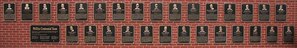 Forty black metal plaques are mounted on a brick wall. They are inscribed with gold images of human faces and text.