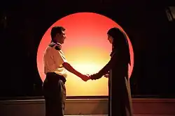 Two actors stand together, holding hands against a backdrop of a glowing sunset