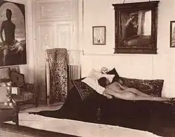 Naked youth poses prone in the Villa Lysis of Capri, where Nino Cesarini lived with his protector and lover, the Baron Jacques d'Adelswärd-Fersen. The photograph was taken by Wilhelm von Plüschow and the portrait of Cesarini is visible on the wall.