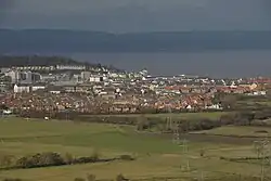 Multiple buildings including terraces, detached houses and blocks of flats. In the foreground are fields and in the background water and then hills.