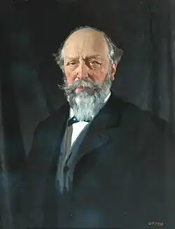 Portrait of a balding middle-age man with grey hair and beard, wearing a dark overcoat over a blue jacket and white shirt.