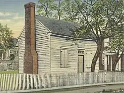 Colorized photo of a log-cabin-style building with a brick chimney and a small tree growing near the door