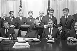 President Lyndon Baines Johnson is sitting with three committee members at a table in the Cabinet Room of the White House. Behind them, nine more committee members are standing, two of them only partially visible.