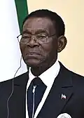 An older man wearing a black suit and tie, appearing stern with short black hair and glasses