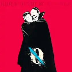 A masked figure holding an unconscious woman in his cloak on a red background. A single blue tear rolls down the woman's cheek. On the man's cloak there is a white circle with a blue line crossed through it. Above them, the words "QUEENS OF THE STONE AGE" and "...LIKE CLOCKWORK" are written in black.