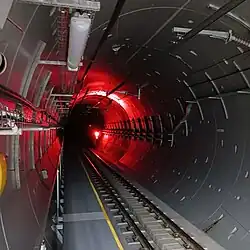 Round concrete tunnel with railway tracks on the bottom, an emergency escape walkway on the left, and various wires and pipes running along the tunnel wall.