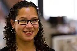 Bracey Sherman smiles directly at the camera. She has light brown skin, glasses, and curly shoulder length hair with the front pulled back.