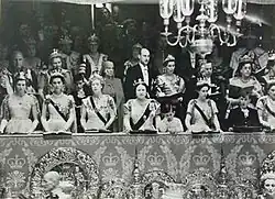 Three rows numerous men and women in fancy dress. Many of the women are wearing tiaras and sashes with white dresses