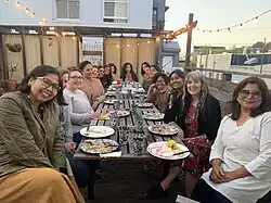 A group of women-identifying and non-binary people sharing a meal and sitting at a long table outside underneath strings of bistro lights at dusk.