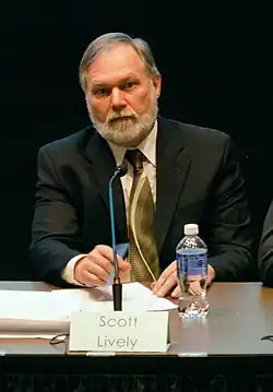 An eldery bearded man with white hair sits in front of a black background. He has a microphone and water bottle. In front of him is a card with his name, reading "Scott Lively".