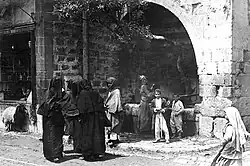 From left to right: A boy staring out from a store's window sill beneath which a lamb is walking by; three fully veiled women conversing on the street; beneath an olive grove jutting out of a large stone archway and beside a fountain, a man is walking, a woman is collecting water from the fountain, and two young boys are standing and smiling; a young girl walking on the street