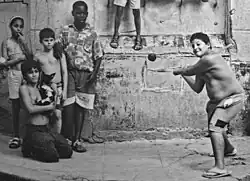 five children stand in-frame and another mostly off-frame; three are wearing no shirts, one of whom is swinging a stick at an airborne ball