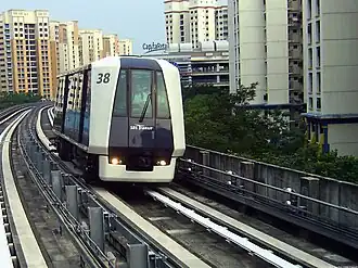 A Mitsubishi Crystal Mover train, numbered 38, on elevated tracks of the Sengkang LRT Line. The train is moving along a curve with multi-story residential buildings in the background.