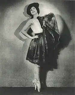 A white woman dancer, posing in toe shoes, one hand on a hip, wearing a hat, a shawl, and a short dark skirt