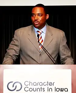 Andre Ware standing a podium in a grey suit. The podium reads "Characters Counts In Iowa".