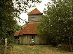 The end of a church seen between trees; the ground floor is timber framed, above this is a weatherboarded tower, and to the left part of the stone body of the church. All the roofs are covered in red tiles